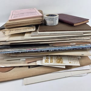 May include: A large stack of assorted paper ephemera, including vintage documents, envelopes, and cardstock. A small roll of decorative tape and a dark maroon rectangular notebook rest on top of the pile, which features various textures and colors.