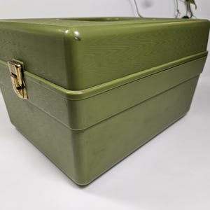 May include: An olive green vintage box with a textured wood grain pattern on the top section. The box has a rectangular shape with a hinged lid and a gold-colored clasp. The box is likely used for storage.