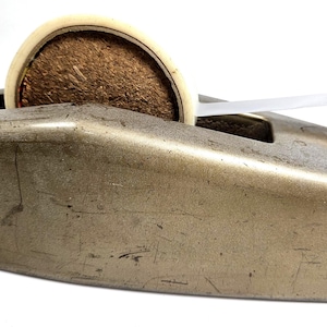 May include: A heavy-duty, metallic gold-toned desktop tape dispenser with a weathered, scratched finish. It holds a roll of tape with a brown, textured core and features a serrated metal cutting edge at the front for dispensing clear adhesive tape.