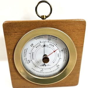 May include: A square wooden barometer with a brass-toned circular face and a hanging ring at the top. The white dial features black text including Stormy, Rain, Fair, and Dry, with a black needle and a red indicator needle pointing toward Dry.