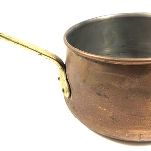 May include: A round copper saucepan with a polished brass handle and a smaller brass side handle. The interior of the pot is lined with a silver-colored metal. The exterior surface of the copper pot shows natural variations in color and texture.