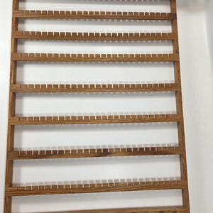 May include: A rectangular wooden jewelry organizer featuring seven horizontal slats with evenly spaced notches for hanging items, and a bottom base rail equipped with ten small, protruding wooden pegs for additional storage and display.