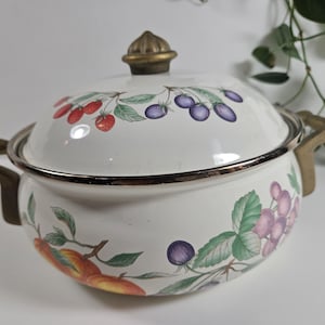 Puede incluir: Olla de esmalte blanco con tapa, con un diseño de frutas de cerezas, ciruelas y uvas. La olla tiene asas de color latón y un pomo a juego en la tapa. Un artículo de menaje de cocina decorativo.