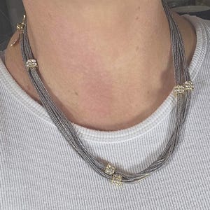 May include: A multi-strand necklace featuring thin, metallic silver-toned wires gathered together. The necklace is accented with several small, cylindrical gold-toned beads encrusted with clear, sparkling crystals spaced along the strands.