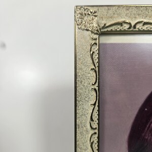 May include: A rectangular, antique-style picture frame with a decorative floral corner design. The frame has a distressed, metallic finish with ornate, curved detailing along the edges. The image inside the frame is partially visible, showing a person.