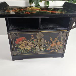 May include: A black wooden cabinet with two doors and a shelf. The doors are decorated with floral designs in gold, red, and green. The cabinet has a black lacquered finish.