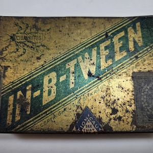 May include: A vintage tin box for In-B-Tween cigarettes. The box is gold with green and black lettering and a green and black insect logo. The text "Copyright 1914" is visible on the box.