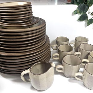 May include: A large stoneware dinnerware set featuring a stack of brown-rimmed plates and bowls alongside several light beige mugs and a matching creamer pitcher. Each piece has a smooth, speckled finish with a distinct dark brown edge.