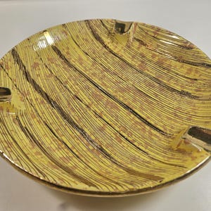May include: Yellow ceramic ashtray with a gold rim and three cigarette rests. The ashtray has a textured surface with a pattern of black and brown lines. A vintage piece for home decor.