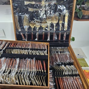 May include: A wooden box with a brass latch, containing a set of 50 gold-tone silverware pieces with wooden handles. The box is labeled "Solid Nickel-Bronze" and "The Only Lifetime Guarantee Genuine Thai Bronze"