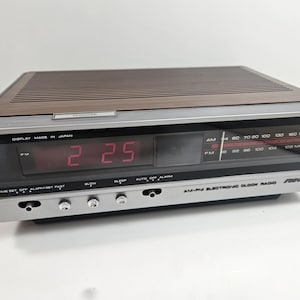 May include: A vintage AM-PM electronic clock radio with a silver and brown design. The digital display shows the time as 2:25. The radio has an AM/FM tuner and several control buttons. The text "DISPLAY MADE IN JAPAN" is visible.