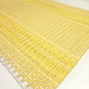 May include: A rectangular woven textile runner featuring a grid pattern of varying shades of yellow. The surface displays a textured, raised weave with horizontal and vertical threads creating a repeating square design across the entire fabric.