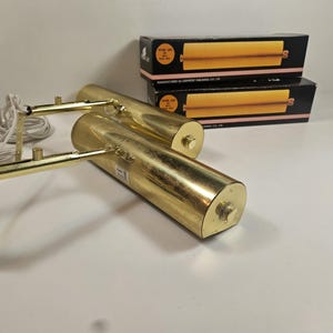 May include: Two gold-colored, cylindrical picture lights with adjustable arms and a power cord. Two boxes are stacked in the background, each with a yellow and black design. The lights are designed for artwork illumination.