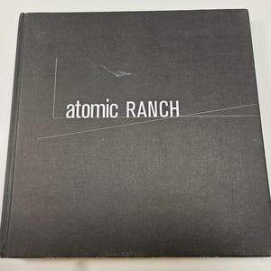May include: A dark gray book with the title "atomic RANCH" in white lettering. The book cover has a textured appearance with a white line design. The book is on a white surface.