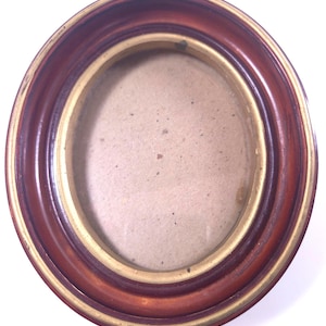May include: An oval picture frame featuring a deep reddish-brown wooden outer border and a thin, metallic gold-toned inner rim. The center contains a plain, light beige backing board, and the entire structure is enclosed by a clear glass pane.