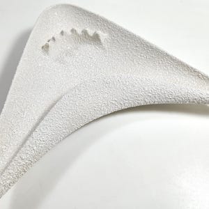 May include: A white, triangular-shaped dish with a textured, granular surface. The object features a distinct, curved, boomerang-like silhouette with a recessed area containing a series of small, molded indentations along one of the inner edges.