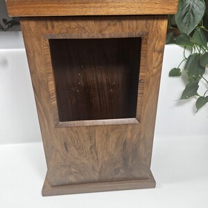 May include: A wooden cabinet with a rectangular opening. The cabinet is made of dark brown wood with a visible wood grain pattern. The top and base are a lighter shade of brown. The cabinet has a trapezoidal shape.
