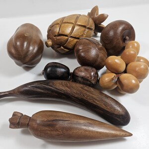 May include: A collection of handcrafted wooden fruit sculptures, including a pineapple, pear, grapes, and a banana. The sculptures are various shades of brown and have detailed carvings, showcasing the natural wood grain.