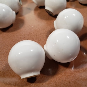 May include: A close-up of ten white ceramic round knobs with a smooth, glossy finish. The knobs are arranged on a brown surface.