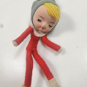 May include: A vintage doll with a fabric body and a painted face. The doll has a red outfit, a gray hat, and yellow hair. The doll's feet and collar are white.