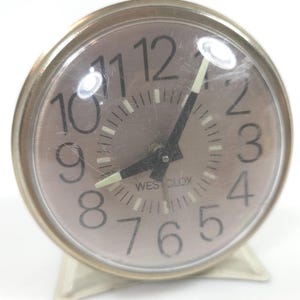 May include: A round Westclox alarm clock with a metallic silver face, black Arabic numerals, and black hands with luminous tips. The clock is encased in a clear, slightly scratched protective cover and features a simple, integrated metal stand base.