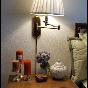 May include: A bedroom scene featuring a wooden nightstand with a wall-mounted lamp and decorative items. The lamp has a pleated shade. The nightstand holds candles, a small box, a white decorative jar, and a vase of purple flowers.