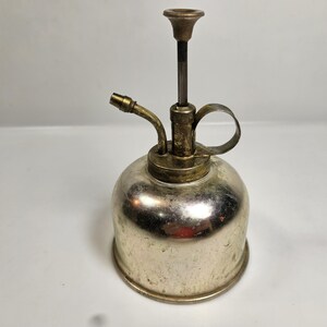 May include: A vintage silver-toned plant mister with brass accents. The mister has a bulbous body, a curved nozzle, and a handle. The top features a brass fitting and a vertical stem with a small, decorative top. The mister is designed for spraying water on plants.