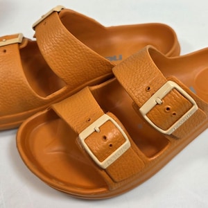 May include: A pair of orange slide sandals featuring two wide straps with a textured finish. Each strap is secured by a light beige adjustable buckle. The footbed is contoured with a smooth surface, and the overall design is molded from a single material.