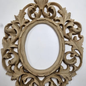 May include: An ornate, oval-shaped picture frame with a beige finish. The frame features elaborate, carved floral and scrollwork designs. The frame has a decorative top element and a small base, suitable for wall mounting. The inner oval is empty.