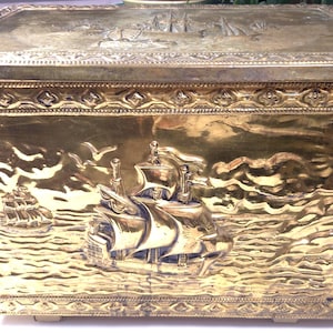 May include: A rectangular brass box featuring an embossed nautical scene on the front and lid. The front panel depicts a large sailing ship on the ocean with smaller vessels in the distance, framed by a decorative border of repeating geometric patterns.