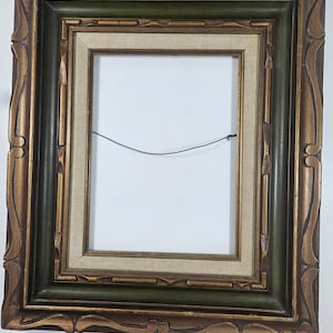 May include: Ornate, rectangular picture frame with a gold and green finish. The frame has a detailed, carved design along the edges and a neutral-colored canvas backing. A thin wire is strung across the back of the frame.