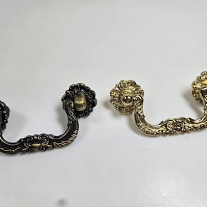 May include: Two ornate drawer pulls. One is dark bronze, the other is gold. Both have a floral design on the ends and a curved handle. The drawer pulls are likely for a cabinet or dresser.