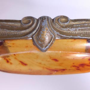 May include: A drawer pull featuring an ornate, weathered brass metal frame with scrolling details and a central, oval-shaped insert made of translucent, mottled amber-colored material with dark brown, marbled streaks throughout the surface.