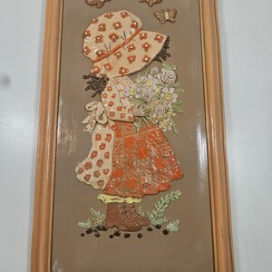 May include: A framed relief sculpture of a girl holding flowers, wearing a bonnet and dress with floral patterns. The artwork features a brown background, a wooden frame, and three butterfly accents.