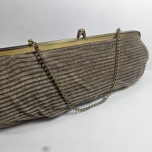 May include: A vintage-style clutch purse in a neutral taupe color. The purse features a corduroy-like texture and a gold-toned metal frame with a chain strap. The clasp is a simple, elegant design.