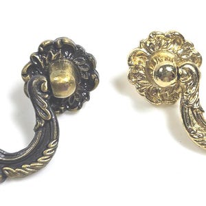 May include: Two metal drawer pulls with ornate, curved handles and circular backplates. One pull features a dark, antique bronze finish, while the other has a bright, polished gold finish. Both handles display intricate, raised floral detailing.