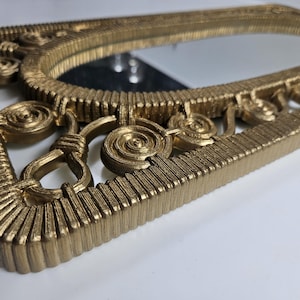 May include: A gold-colored, rectangular mirror with an ornate frame. The frame features a textured, ridged design and decorative spiral and loop accents. The mirror's surface reflects the surrounding environment.