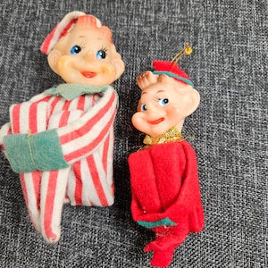 May include: Two vintage Christmas elf ornaments. One elf is wearing a red and white striped pajama-like outfit with a green hat. The other elf is wearing a red felt outfit with a green hat and a gold trim.