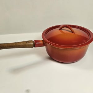 May include: A red-orange saucepan with a lid and a wooden handle. The handle is attached to the side of the pan with a metal ring. The pan is on a white surface.