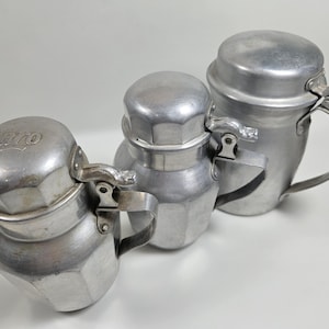 May include: Three silver aluminum pitchers with lids and handles. The pitcher on the left has the word "Karo" embossed on the lid.