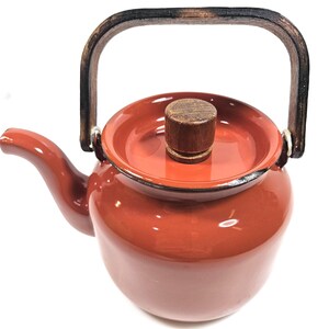May include: A glossy terracotta-colored teapot featuring a rounded body, a curved spout, and a matching lid with a cylindrical wooden knob. The teapot includes a dark, weathered-looking arched wooden handle attached with silver-toned metal hardware.