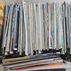 May include: A large collection of folded paper maps in a clear plastic storage bin. The maps are organized in rows and columns, with some maps sticking out from the bin. The maps are mostly in shades of white, gray, and blue.