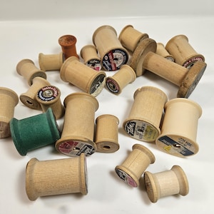 May include: A collection of vintage wooden spools in various sizes and colors. Some spools have labels with text and designs. One spool is green, and another is orange. The spools are scattered on a white surface.