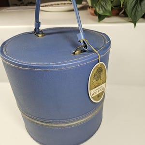 May include: A vintage blue wig case with a handle and zipper closure. The cylindrical case has a stitched design and a tag that reads "the miner wig case". The handle is attached with gold-colored hardware.