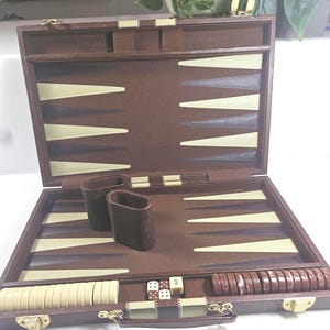 May include: A brown backgammon set in a briefcase-style case with a felt interior. The board features cream and dark brown triangular points. Included are two brown leather dice cups, a set of cream and brown checkers, and four standard dice.