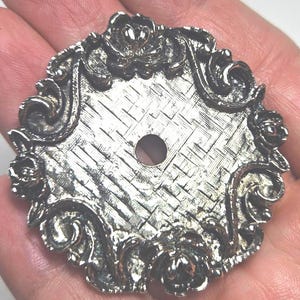 May include: A circular silver-toned metal furniture plate featuring a central hole and a textured basket-weave pattern in the middle. The outer edge is decorated with a raised relief design of floral motifs and scrolling curves in a metallic finish.