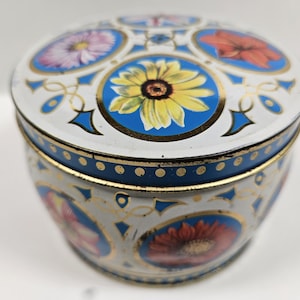 May include: A round, vintage tin with a white base, adorned with blue and gold floral designs. The lid features a central sunflower and other flowers in blue circles. The sides display additional floral patterns, creating a decorative storage container.
