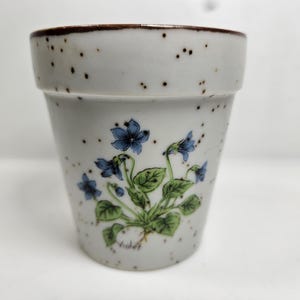 May include: A ceramic flower pot with a speckled, light gray finish and a brown rim. The pot features a floral design of blue flowers and green leaves. The pot is a small size, suitable for indoor plants.