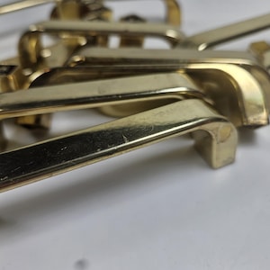 May include: A collection of gold-colored cabinet handles. The handles have a curved design with a rectangular base and a rounded grip. The hardware is made of metal and appears to be vintage or antique.