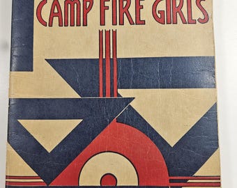 May include: Vintage book cover with the text "BOOK OF THE CAMP FIRE GIRLS." The cover features a geometric design in navy blue, red, and tan. The book appears to be an older edition, with signs of wear.
